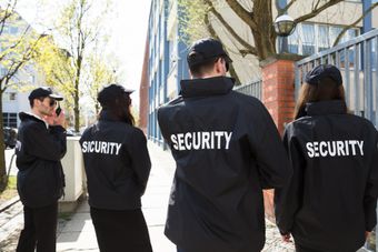 Four security guards in black jackets walk outside a building, seen from behind.
