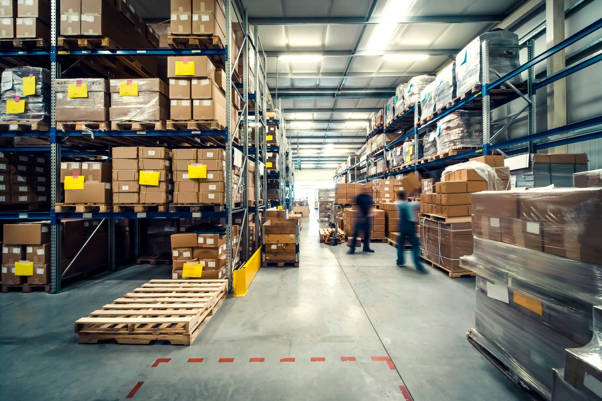 Warehouse aisle with stacked pallets, boxes on shelves, and workers near the center aisle