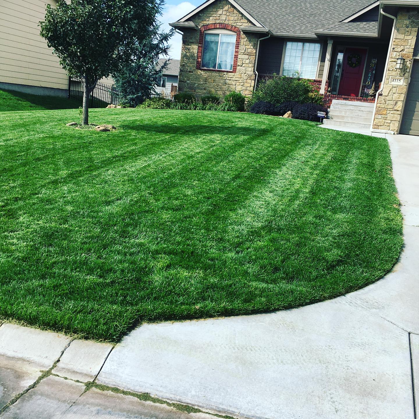 Lush green lawn with striped mowing pattern in front of a house with stone accents and a red door.