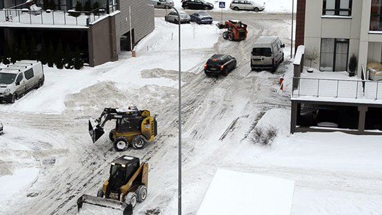 Snow removal equipment clearing a snow-covered street with cars and buildings in the background.