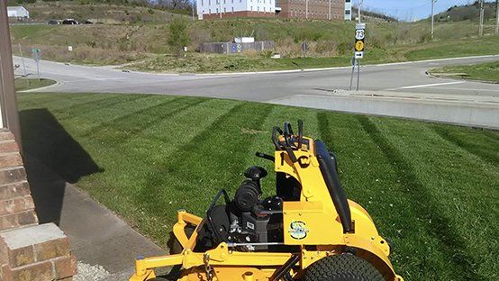 Yellow riding lawnmower mowing grass, creating striped pattern. Near a road and building.