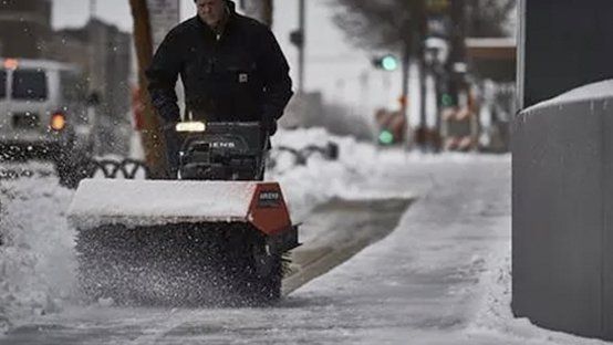 A person using a snow blower on a sidewalk, clearing snow. Urban setting.