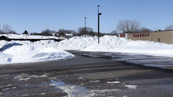 Snow-covered parking lot with large snow piles, partially melted asphalt, and a building with a