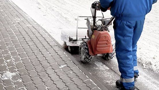 Person in blue jumpsuit using a snowblower on a brick sidewalk. Snow is piled to the side.