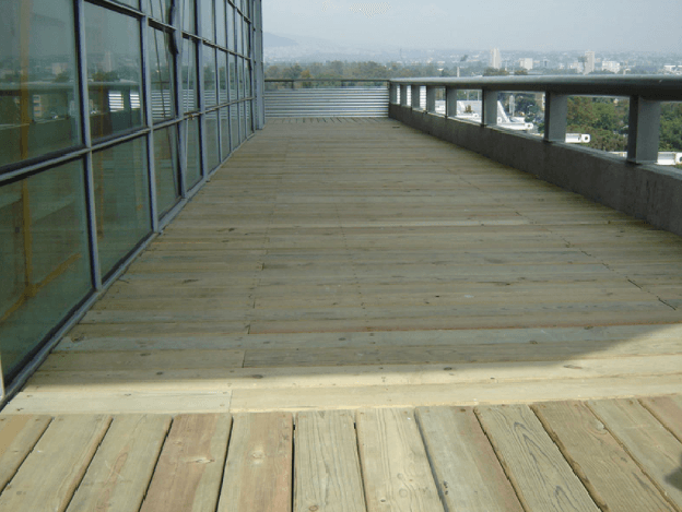 Terraza de madera en la azotea, junto a ventanales y un muro bajo, con vistas al horizonte de la ciudad a lo lejos.