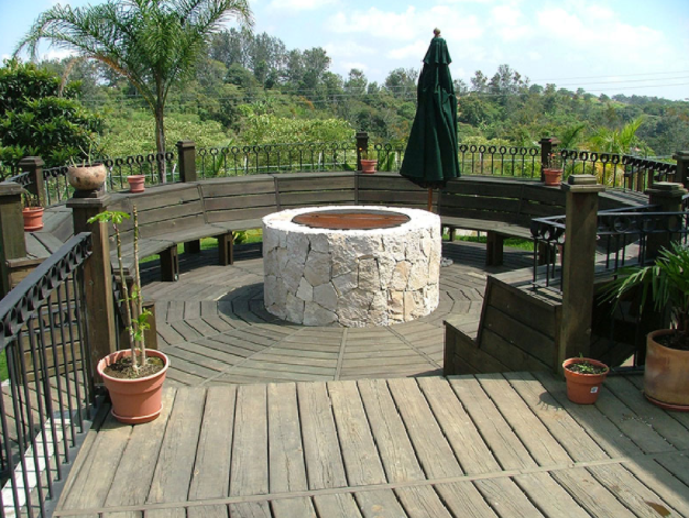 Terraza de madera con bancos curvos alrededor de una hoguera de piedra, con vistas a un jardín exuberante.