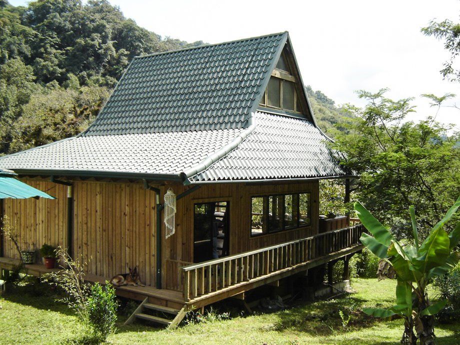 Pequeña casa de madera con techo de tejas verdes, rodeada de exuberante vegetación tropical.