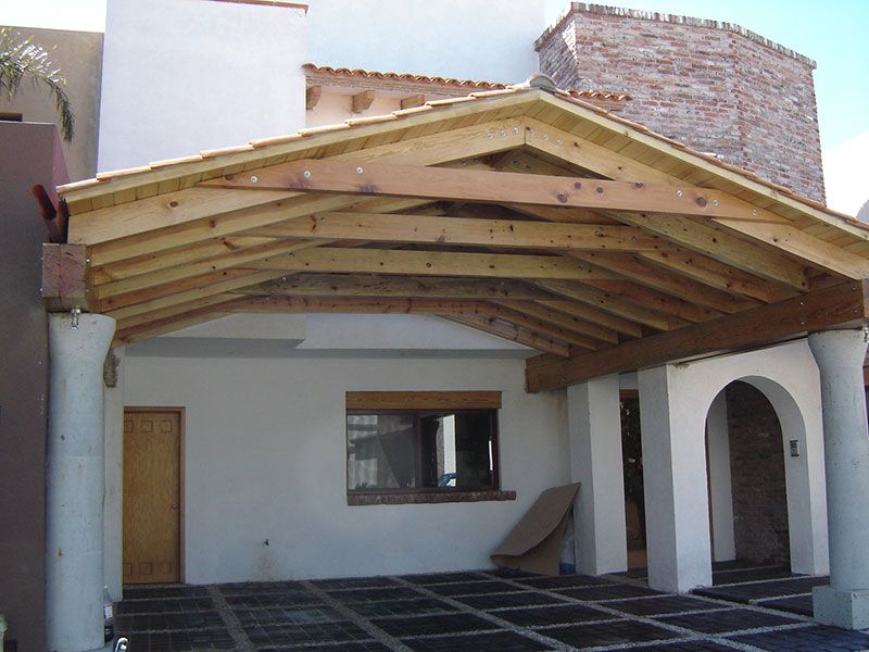 Patio cubierto con vigas de madera en el techo, paredes de estuco blanco, puerta arqueada y suelo de baldosas.