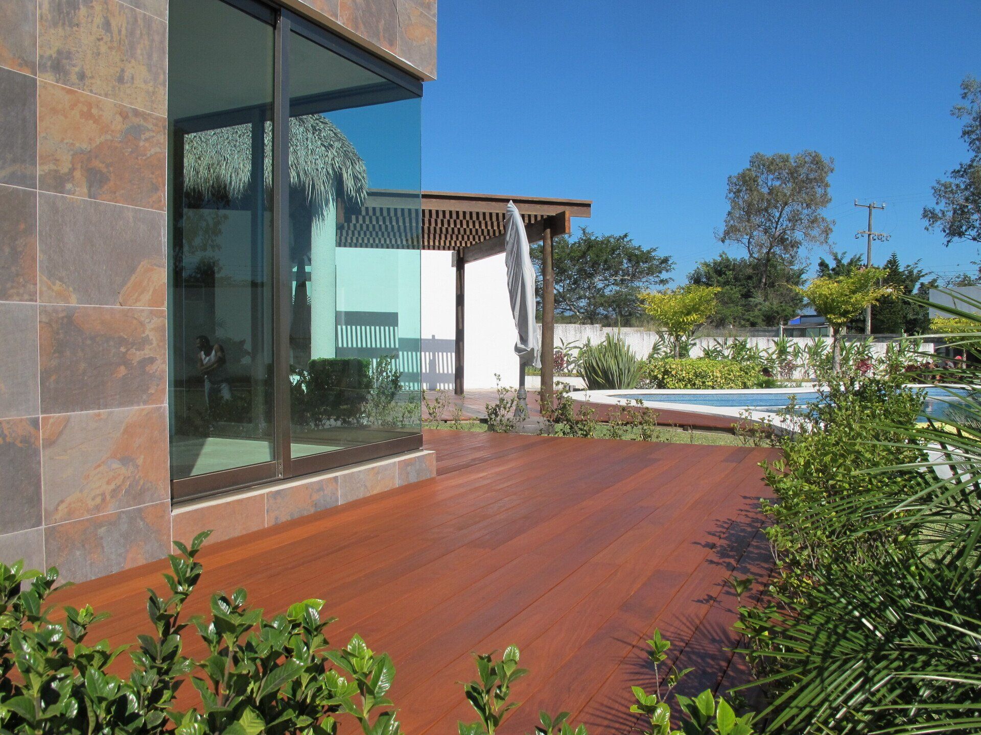 Patio de casa moderna con terraza de madera rojiza, pared de cristal y vegetación junto a la piscina bajo un cielo azul.