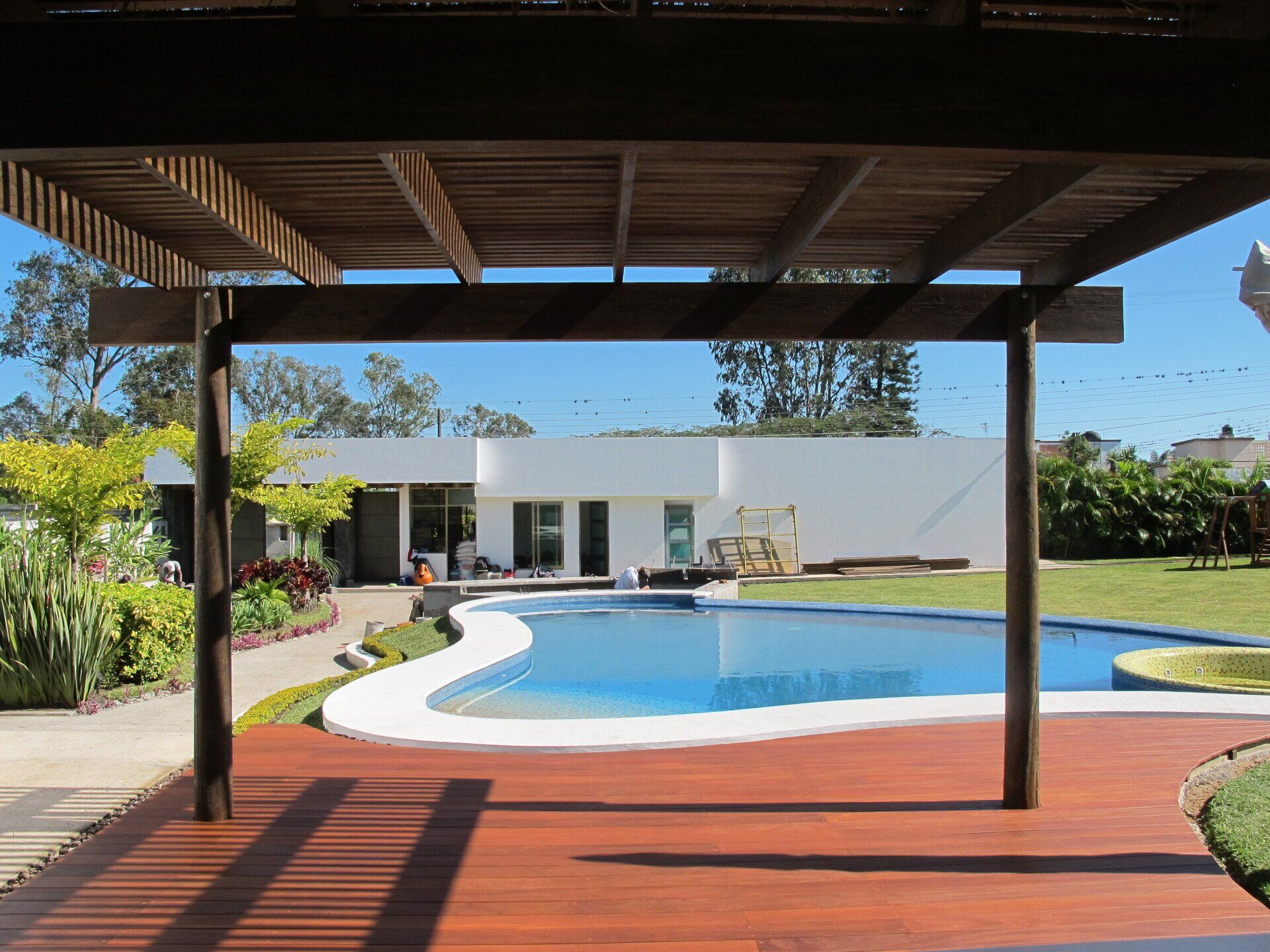 Piscina en el patio trasero con terraza roja, casa blanca y pérgola sombreada en primer plano.