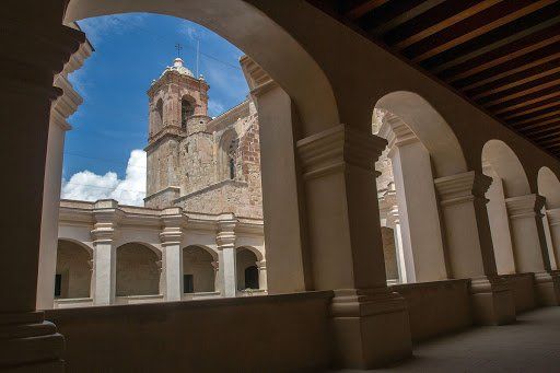 Claustro abovedado que enmarca una torre de iglesia de piedra bajo un cielo azul, con columnas y un pasillo sombreado.