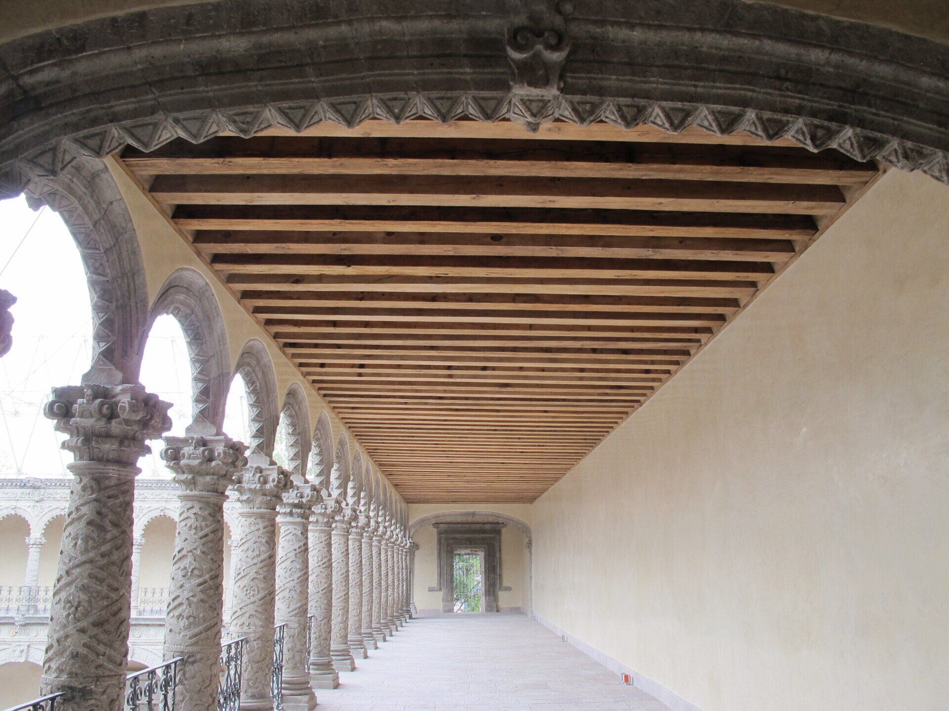 Claustro de piedra cubierto con columnas arqueadas, techo de madera y un largo pasillo vacío bañado por una luz tenue.