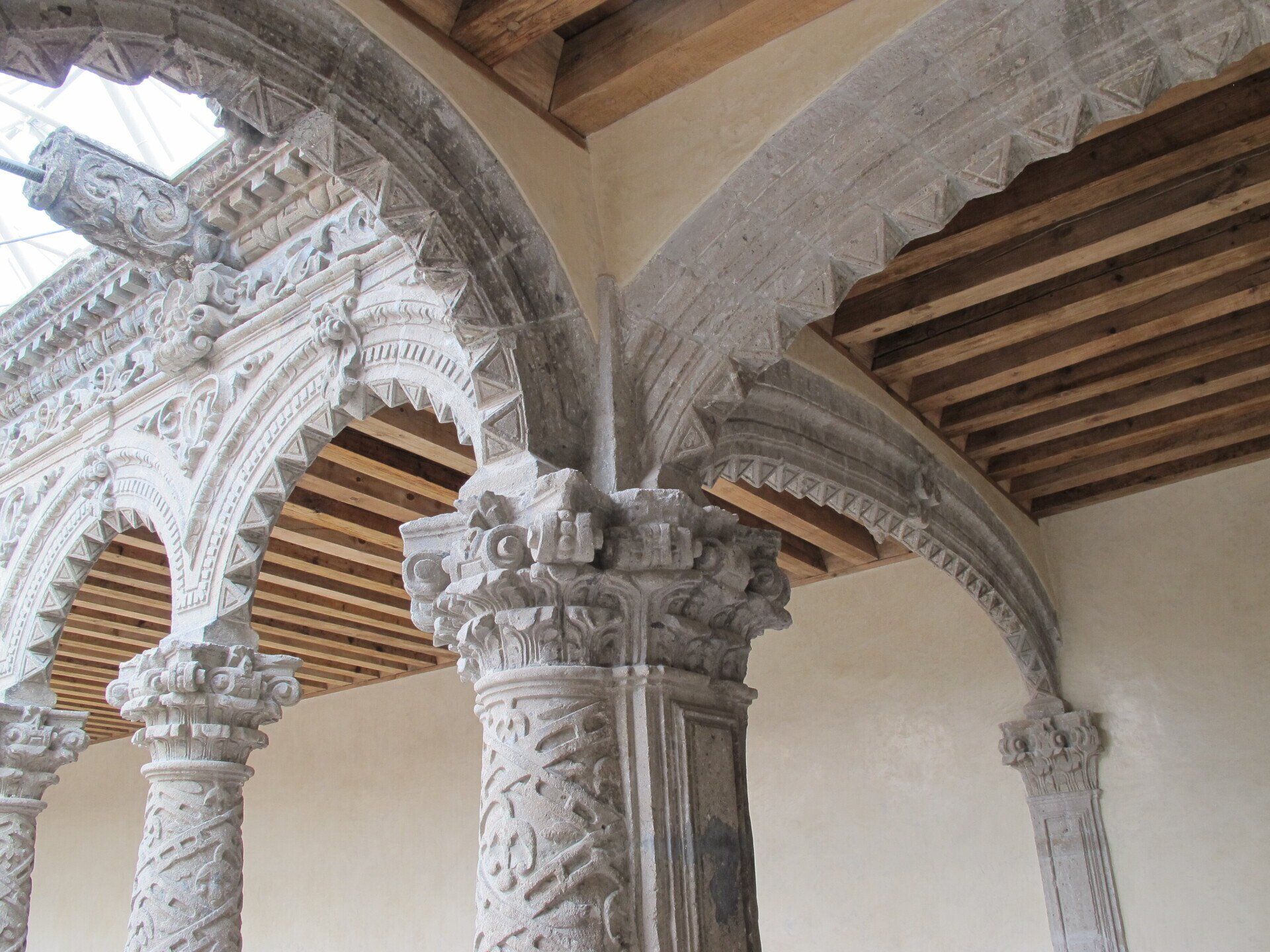 Arcos de piedra ornamentados y columnas talladas en un interior histórico con vigas de madera en el techo.