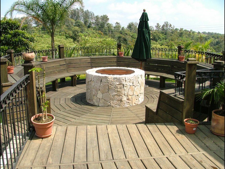 Terraza de madera con una hoguera circular de piedra, bancos curvos, plantas en macetas y una sombrilla de patio cerrada.