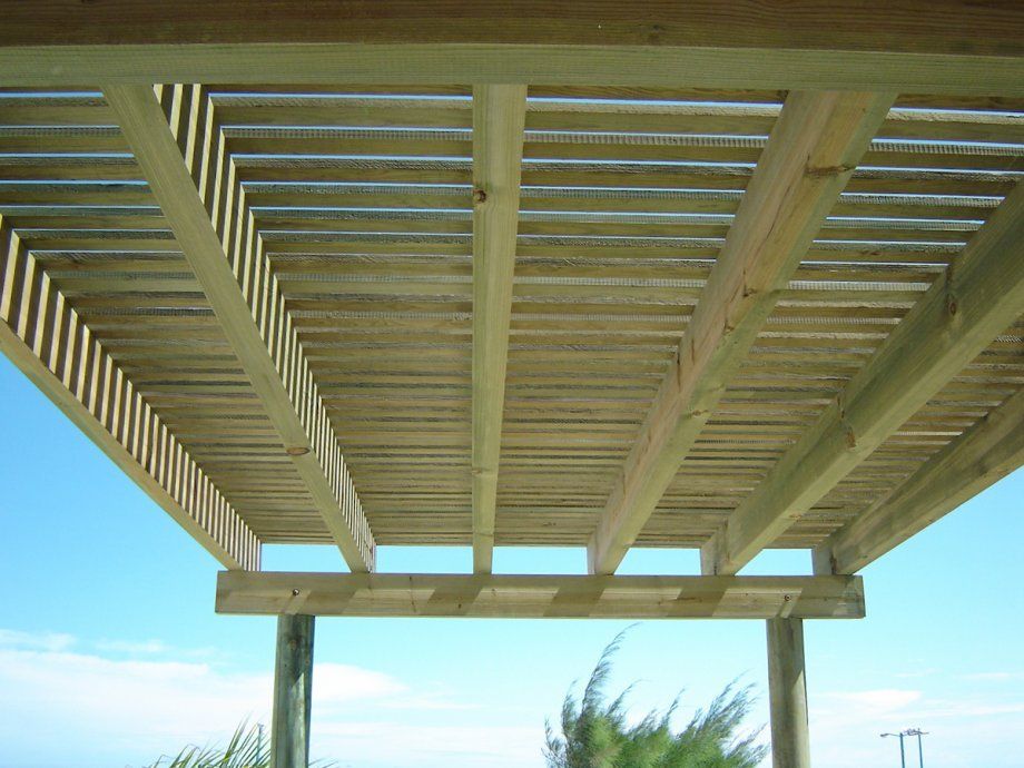 Techo de pérgola de madera visto desde abajo contra un cielo azul y palmeras.