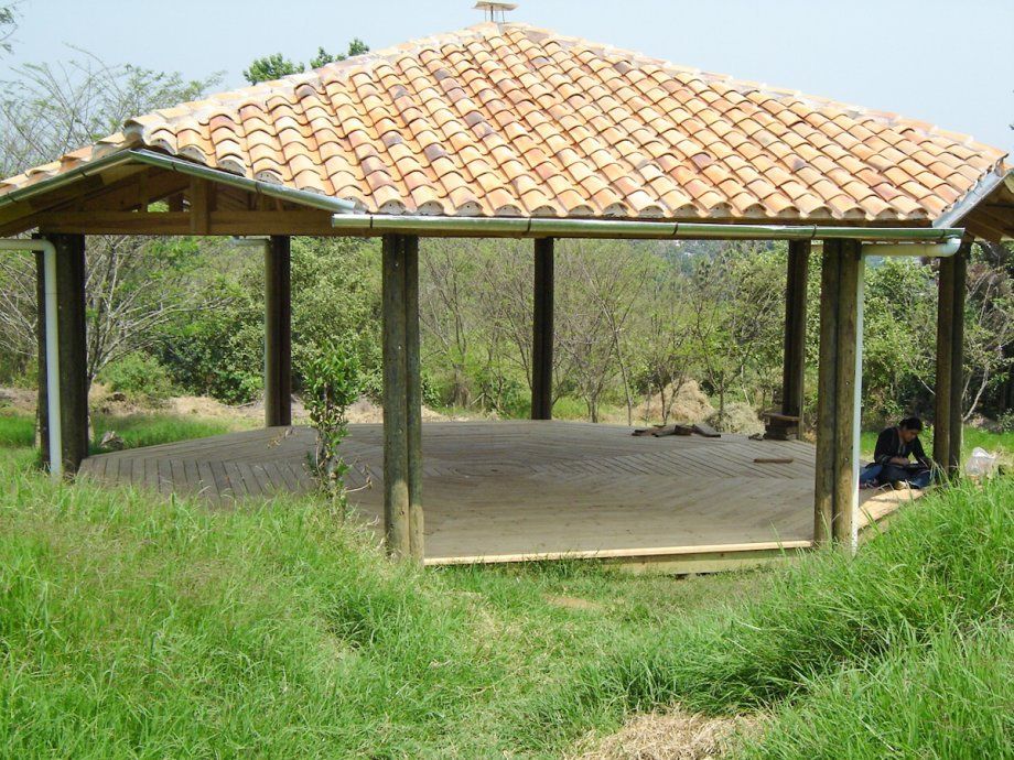 Pabellón abierto por los lados con techo de tejas en una colina cubierta de hierba, rodeado de árboles.