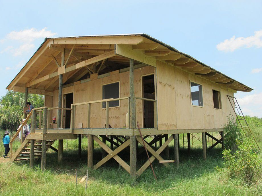 Casa de madera elevada en construcción en un campo de hierba, con una escalera y ventanas abiertas.