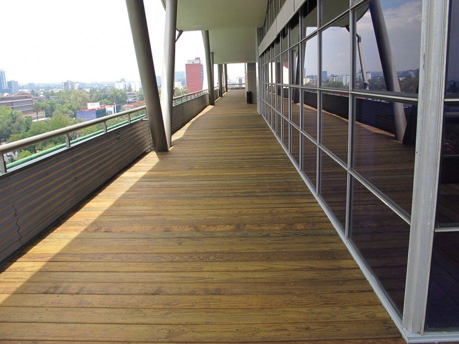 Pasarela de madera en un balcón junto a ventanales, con vistas al perfil urbano de la ciudad.
