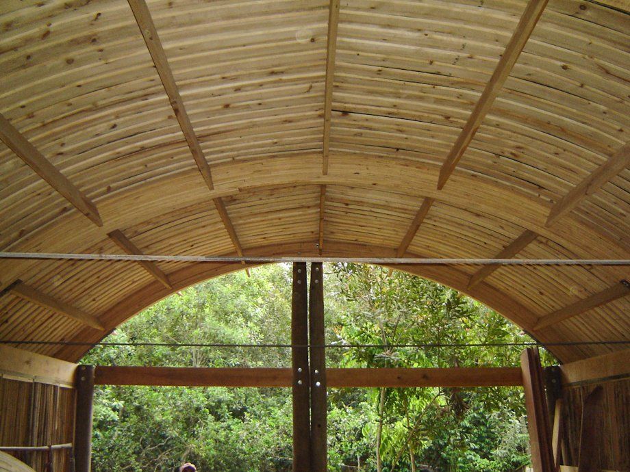 Refugio con techo arqueado de madera que enmarca una vista de un frondoso bosque verde a través de sus lados abiertos.