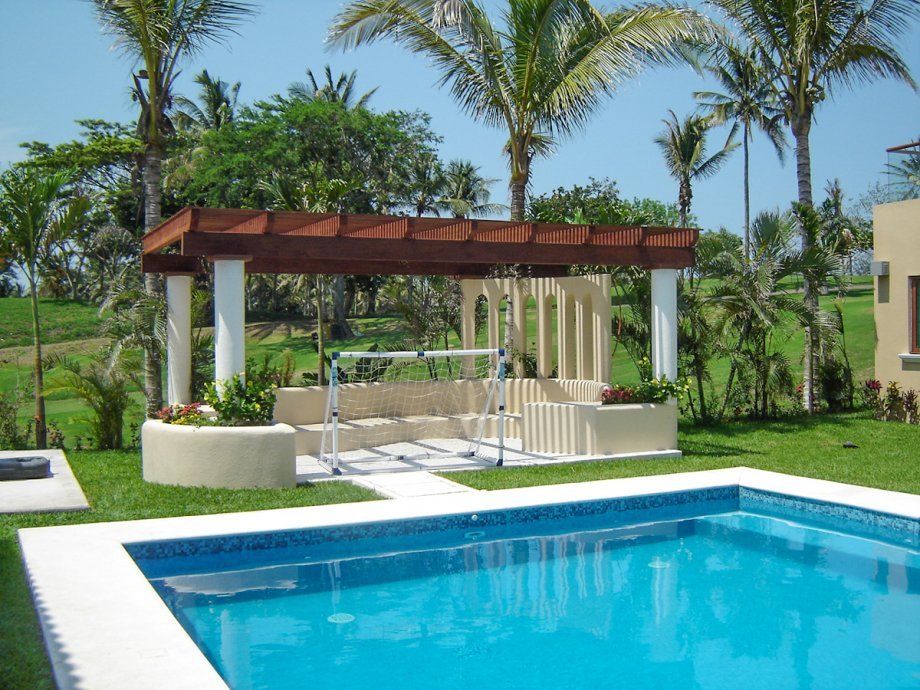 Pérgola junto a la piscina con tumbonas al lado de una piscina azul y palmeras.