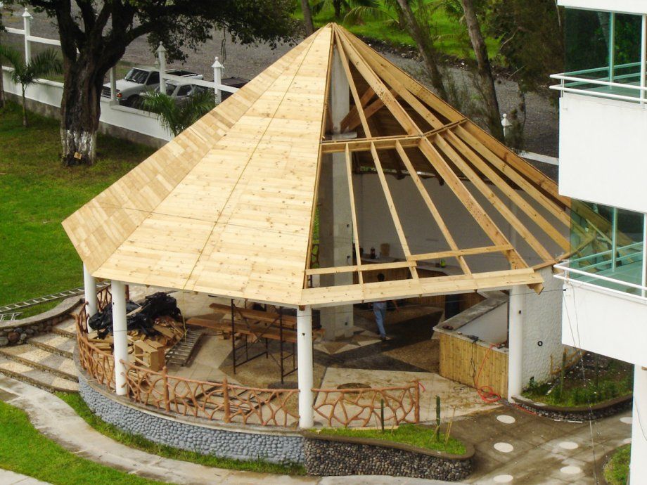 Pabellón abierto por los lados con techo cónico de madera junto a un edificio moderno y árboles.