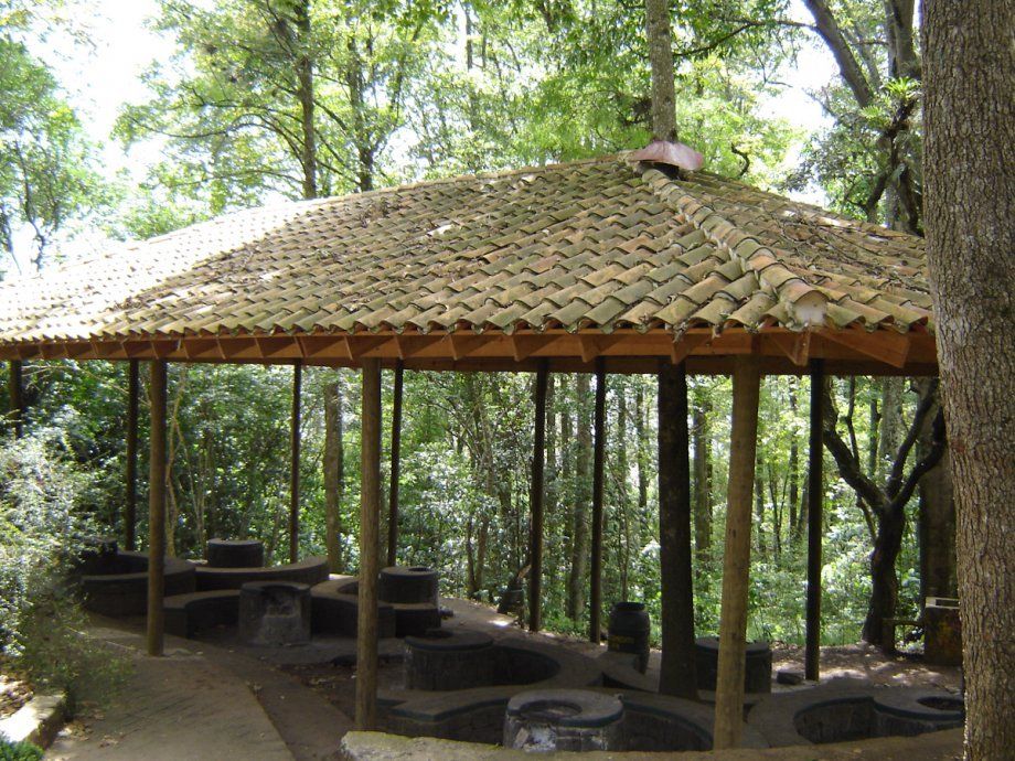 Pabellón rústico al aire libre con techo de tejas, rodeado de árboles en una zona boscosa.