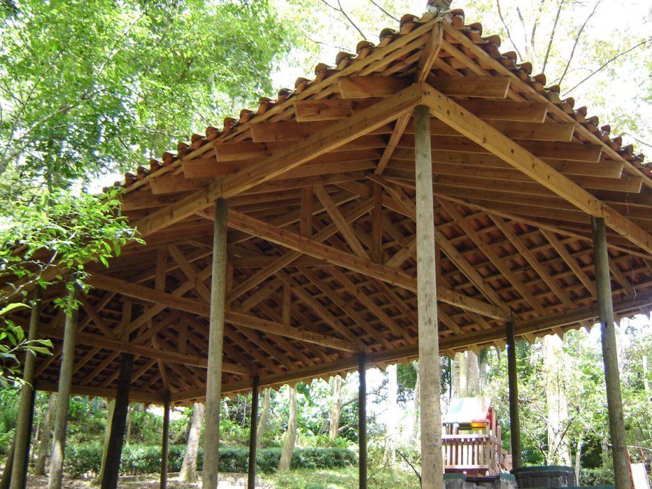Pabellón de madera abierto con techo inclinado en un entorno de parque arbolado.