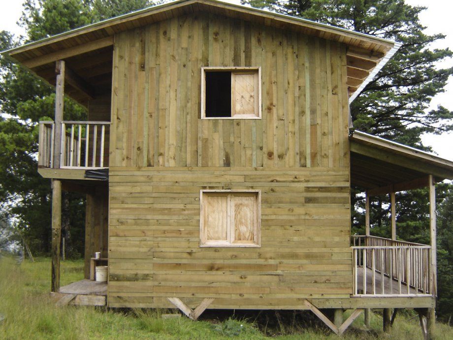 Pequeña cabaña rústica de madera con techo inclinado y dos porches, rodeada de árboles.