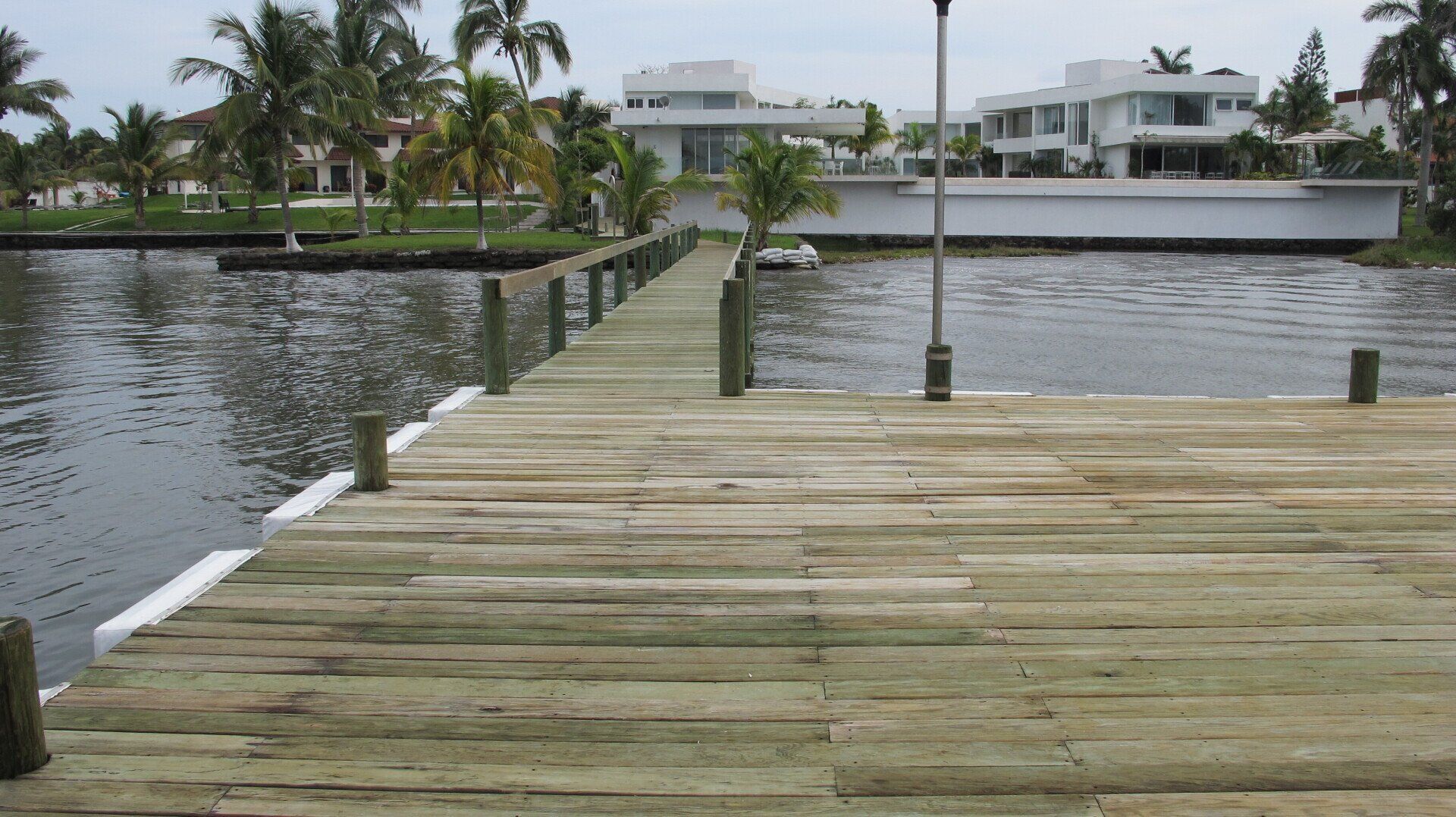 Muelle de madera que cruza el agua hacia modernas casas blancas frente al mar bajo cielos nublados.