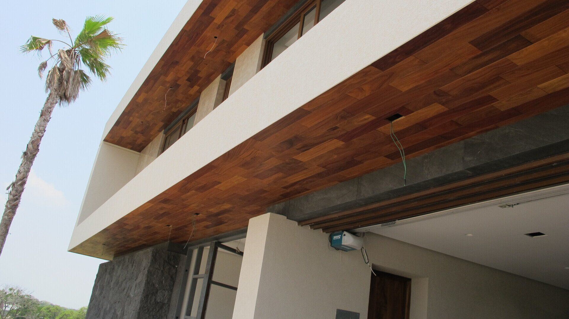 Exterior de una casa moderna con aleros de madera y una palmera alta contra el cielo.