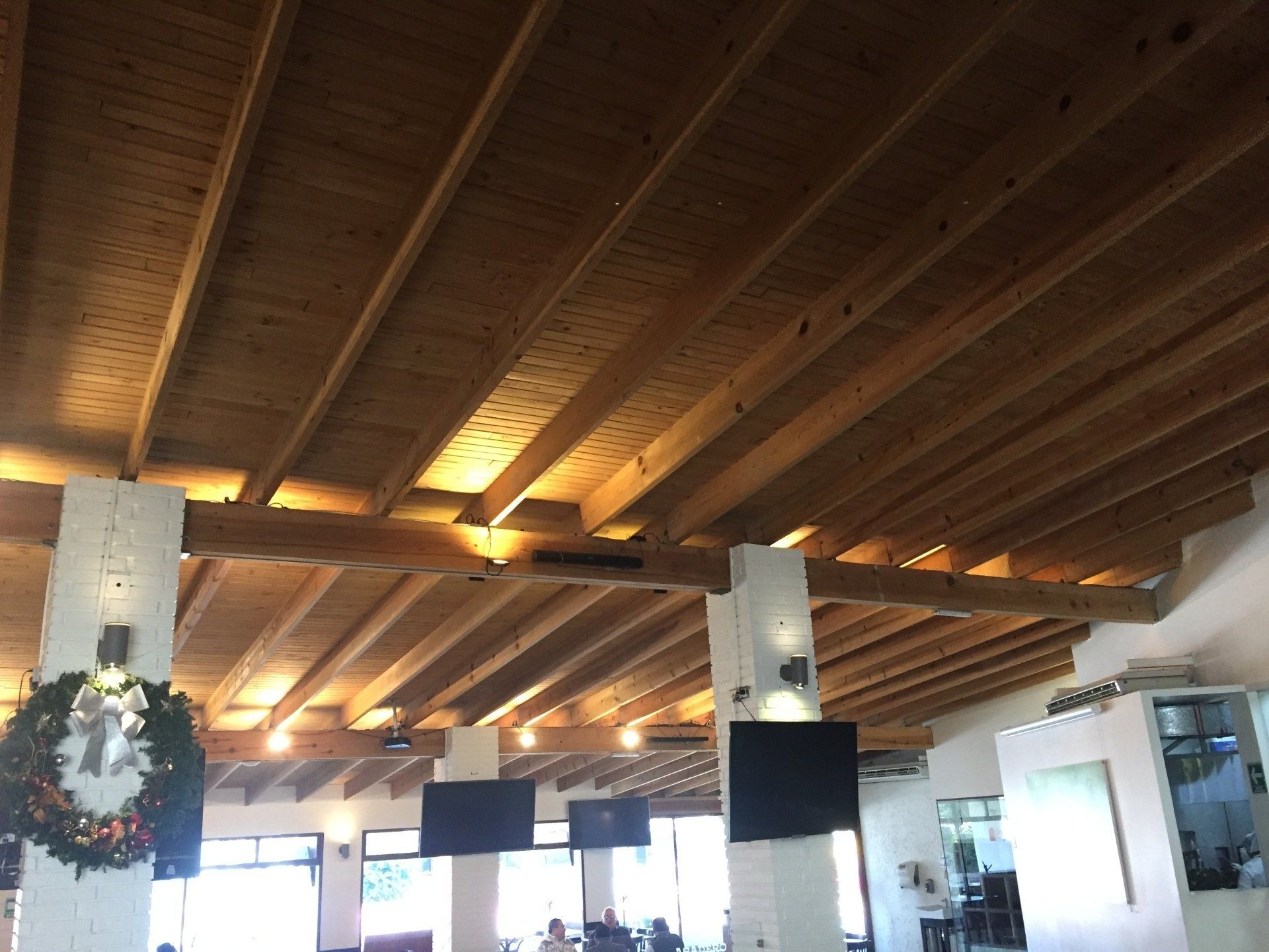 Techo con vigas de madera en una habitación luminosa con luces colgantes y una corona navideña en una columna.