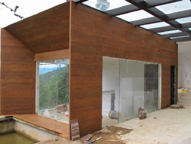 Exterior de una casa moderna revestida de madera en construcción, con grandes ventanales y suelo de hormigón sin acabado.
