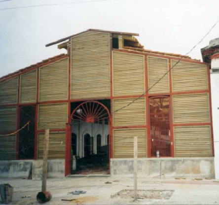 Vista frontal de un pequeño edificio con revestimiento metálico color canela, detalles en rojo y una puerta central arqueada.