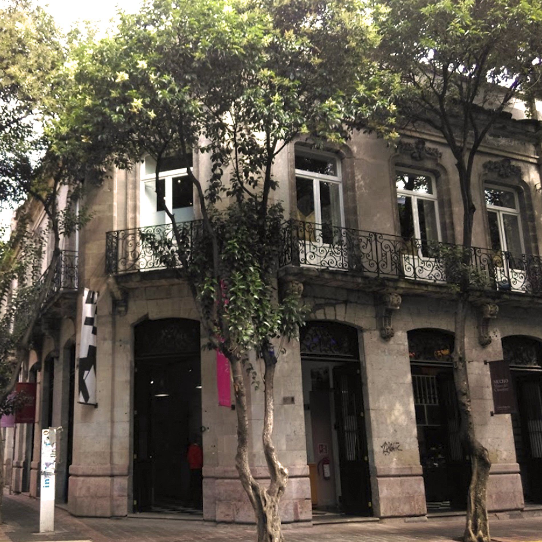 Calle arbolada con un edificio beige de dos plantas y balcones de hierro forjado negro en la esquina.