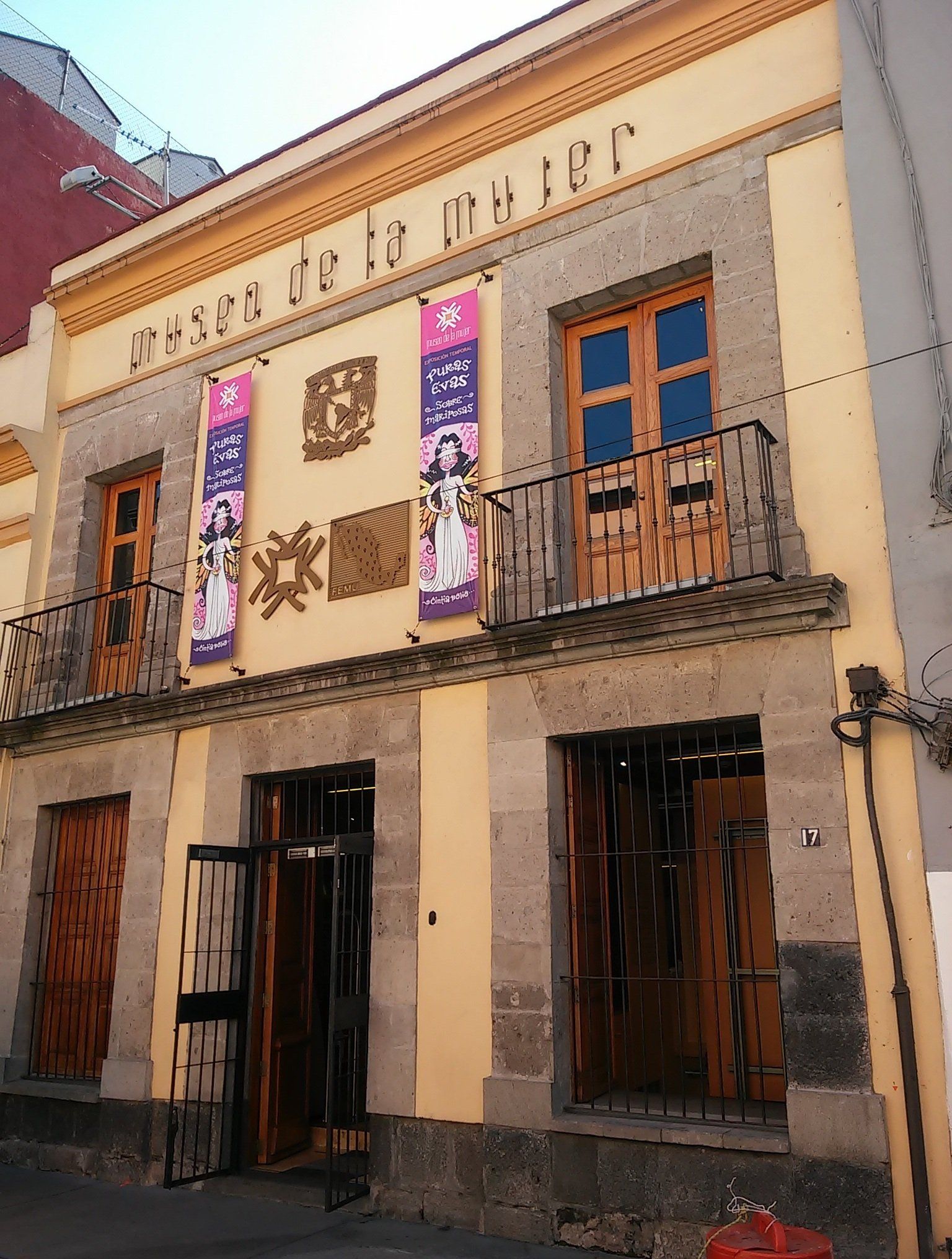Edificio beige de dos plantas con balcones, puertas de madera oscura y pancartas decorativas en las paredes, situado en una calle estrecha.