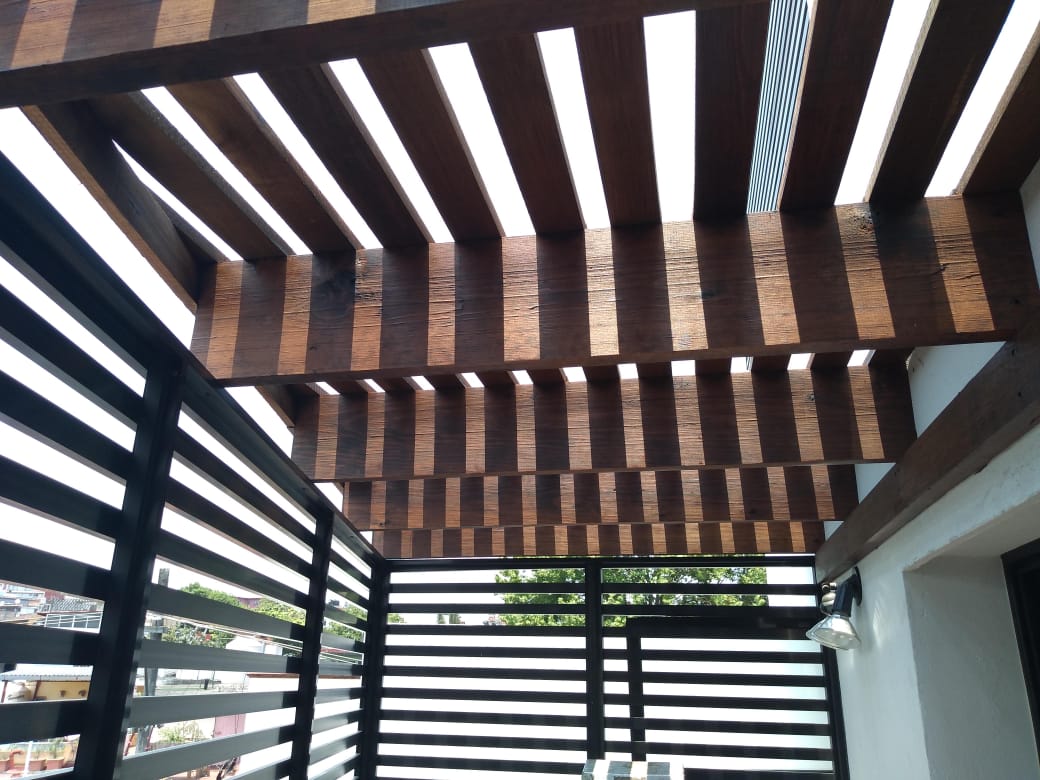 Pérgola de madera con listones a rayas que proyectan sombras sobre un patio y barandilla modernos.