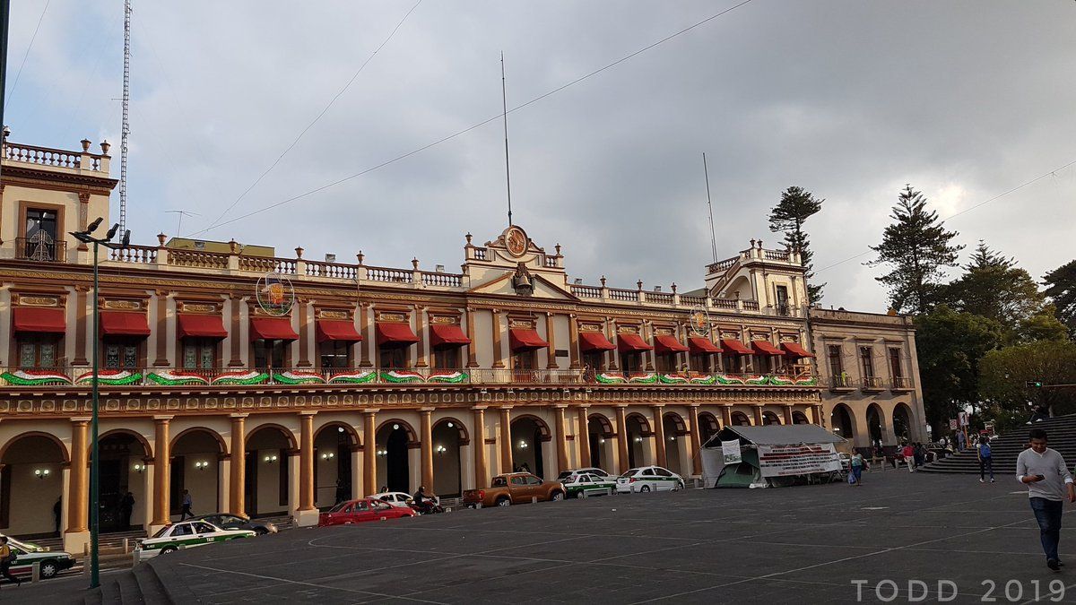 Edificio histórico con arcos y toldos rojos en una plaza bajo un cielo nublado.