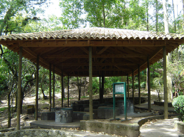Pabellón al aire libre con techo de tejas en un parque arbolado, con bancos de piedra y una bomba de agua central.