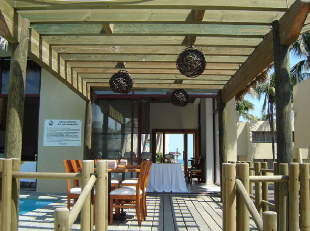 Comedor exterior cubierto con sillas de madera, mesas y faroles colgantes junto a la playa.