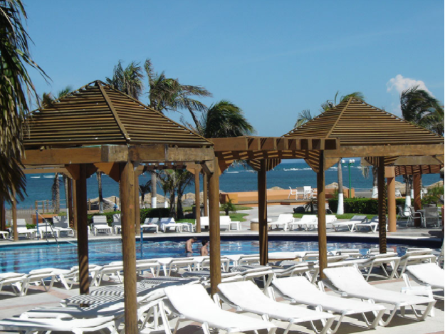 Piscina de un complejo turístico de playa con tumbonas blancas, cabañas de madera y vistas al océano.