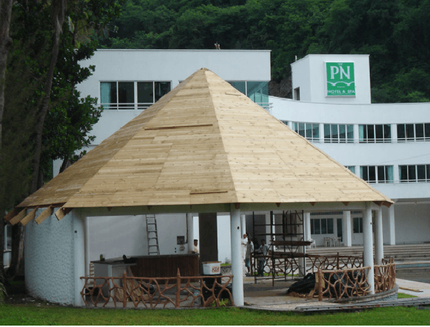 Pabellón circular con techo cónico de madera frente a un edificio blanco y árboles verdes.