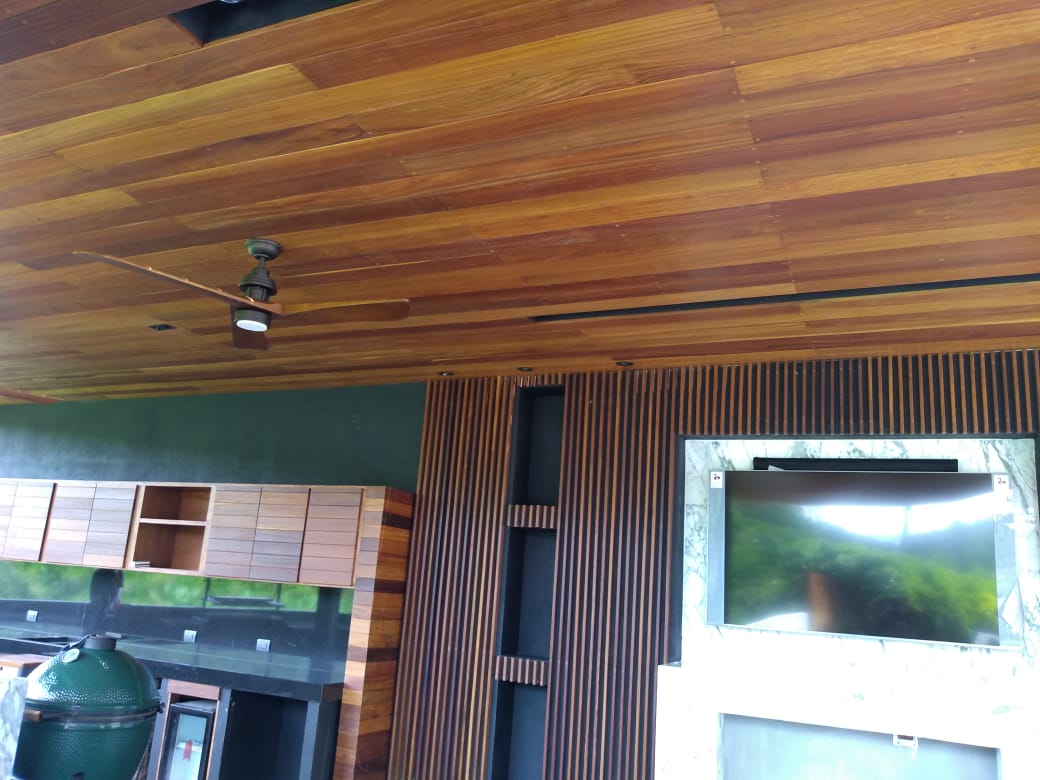 Techo interior con tablones de madera, un ventilador de techo y una pared verde con ventanas y listones de madera.