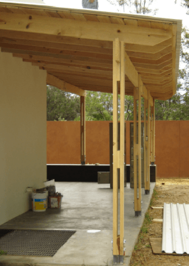 Porche de madera con techo inclinado y postes de soporte junto a una casa, con herramientas y materiales en el suelo.