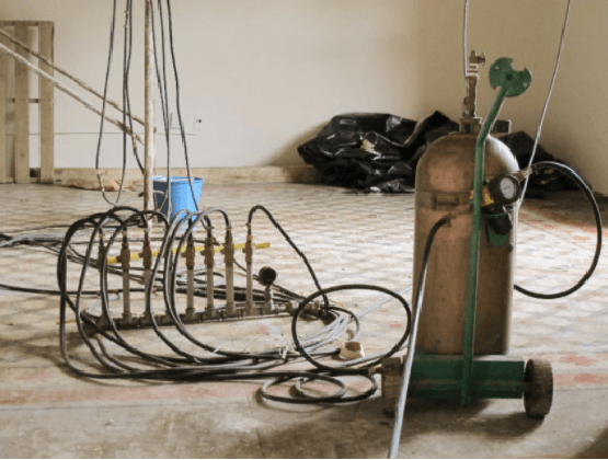 Tubos metálicos enrollados y una bombona de gas verde sobre un suelo de baldosas, con herramientas en una habitación que parece un taller.