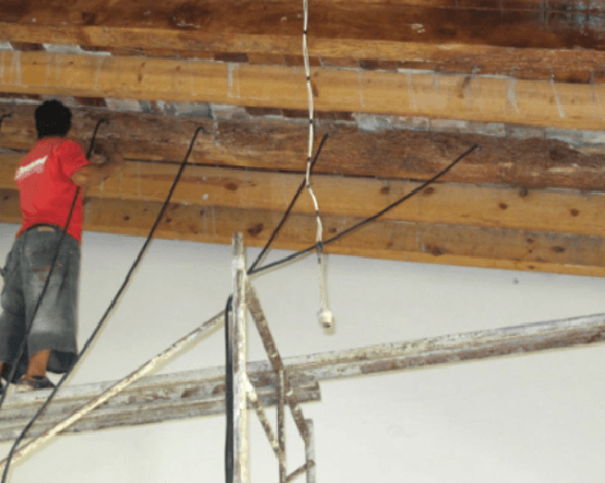 Un trabajador con camisa roja repara el cableado expuesto en una viga del techo, encima de un andamio.