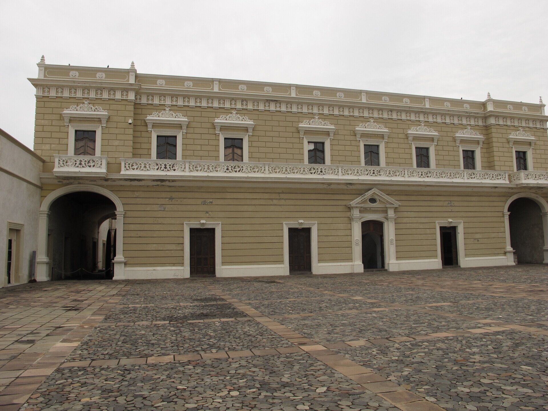 Edificio histórico de piedra beige con entradas arqueadas y patio empedrado bajo un cielo nublado.
