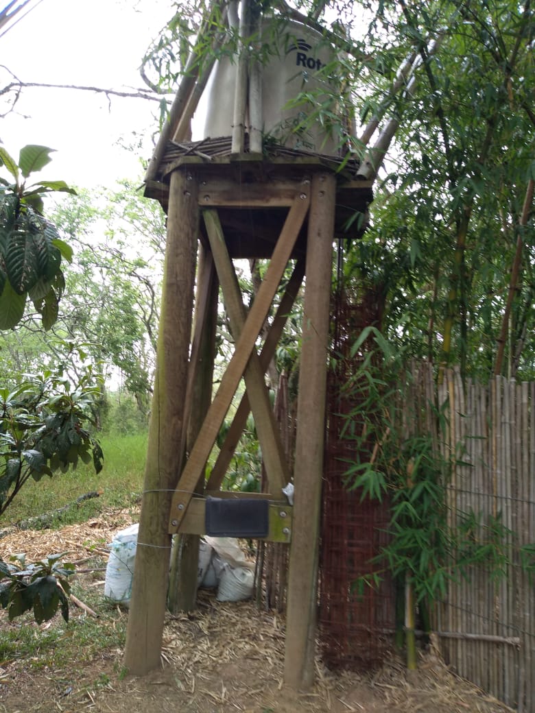 Tanque de agua metálico elevado sobre pilotes de madera junto a árboles en un patio rural.