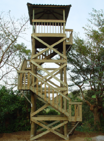 Torre de observación de madera con escaleras y techo, rodeada de árboles y arbustos.