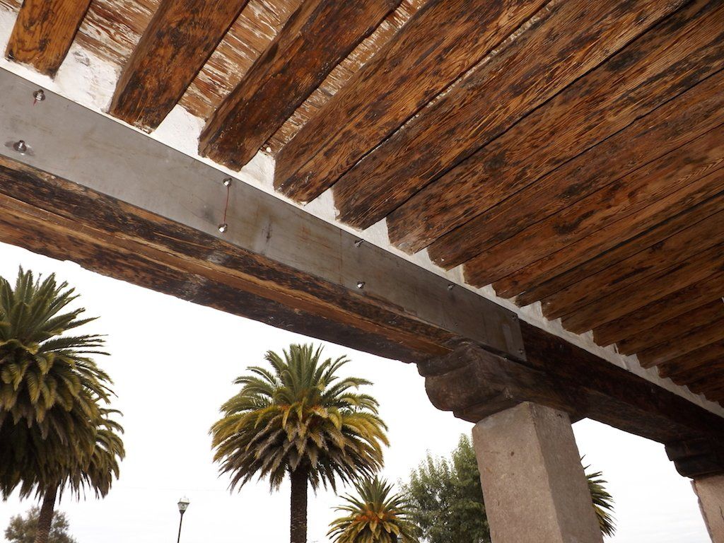 Palmeras enmarcadas bajo una pérgola de madera con un pilar de hormigón contra un cielo brillante.