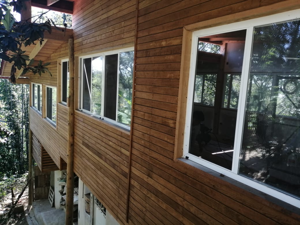 Exterior de una casa revestida de madera con grandes ventanales, situada en una ladera inclinada.